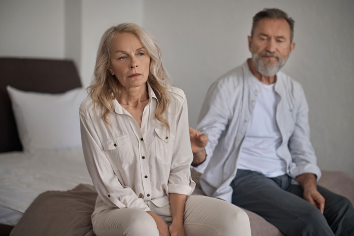 Older couple sitting on bed looking upset, woman avoiding man reaching out, symbolizing breaking up over photograph. Older couple sitting on bed looking upset, woman avoiding man reaching out, symbolizing breaking up over photograph.