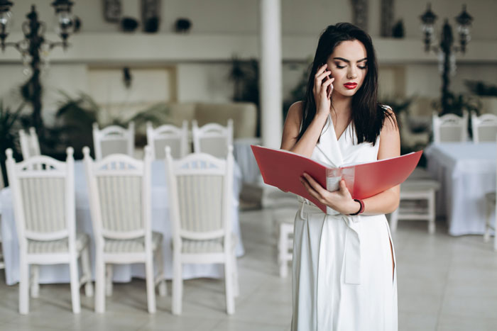 Bride in white dress on phone, reviewing wedding seating chart in a decorated room with white chairs and tables. Bride in white dress on phone, reviewing wedding seating chart in a decorated room with white chairs and tables.