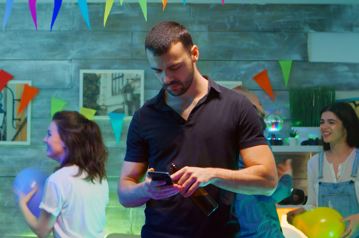 Man at party distracted by phone, holding a beer while others celebrate, reflecting last-minute plan ditch and relationship tension. Man at party distracted by phone, holding a beer while others celebrate, reflecting last-minute plan ditch and relationship tension.