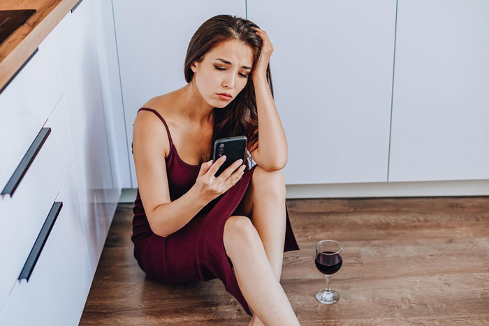 Sad woman in a burgundy dress sits on the floor with phone and a glass of wine after boyfriend ditches plans last minute. Sad woman in a burgundy dress sits on the floor with phone and a glass of wine after boyfriend ditches plans last minute.
