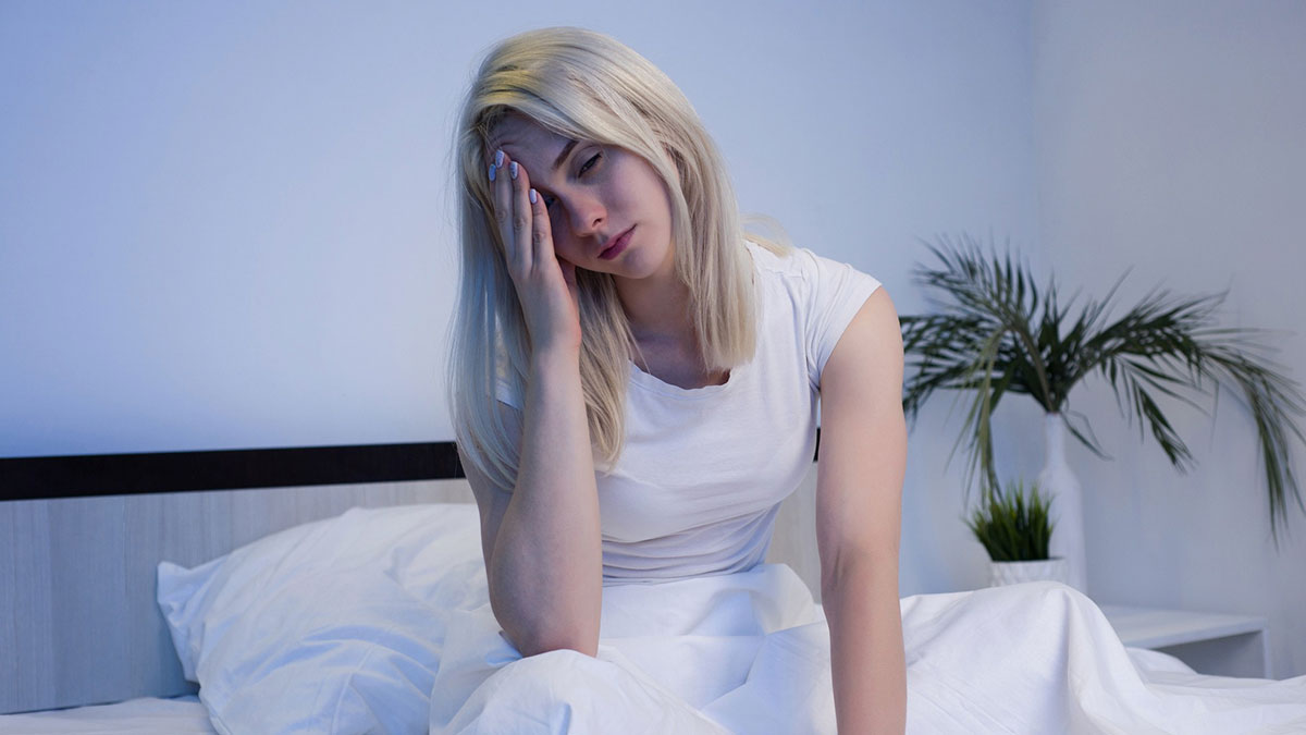 Woman sitting on bed looking tired and stressed, representing heavy periods and anemia symptoms at night.