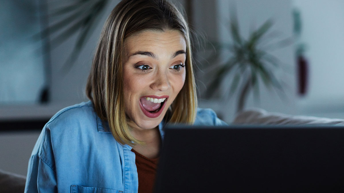 Young woman in casual clothes reacting with surprise to brutal business comebacks on her laptop screen at home.