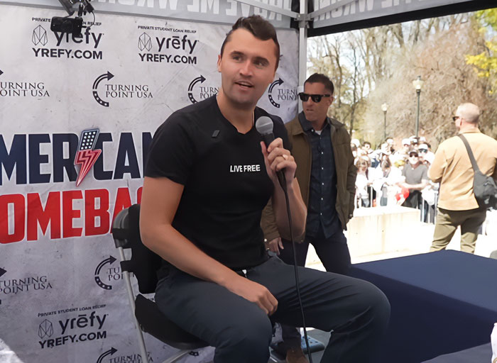 Charlie Kirk speaking at an event, holding a microphone, with a banner showing Turning Point USA and American comeback logos. Charlie Kirk speaking at an event, holding a microphone, with a banner showing Turning Point USA and American comeback logos.