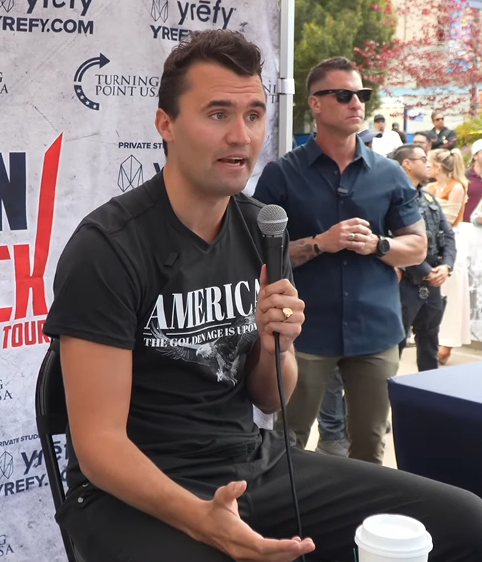 Man speaking into a microphone at a public event with crowd and security in background, discussing Charlie Kirk incident. Man speaking into a microphone at a public event with crowd and security in background, discussing Charlie Kirk incident.