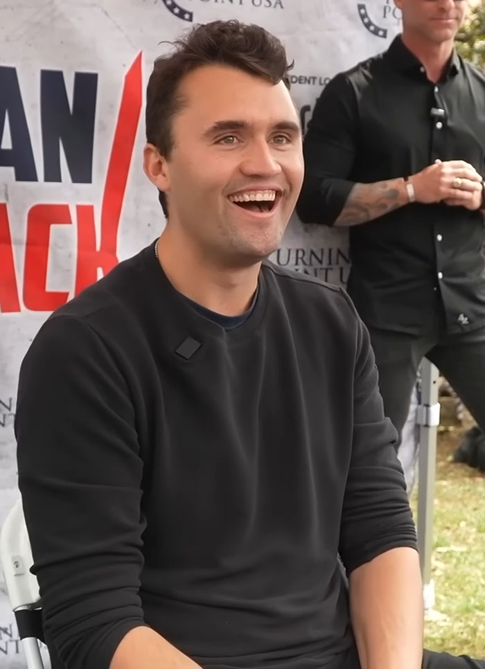 Charlie Kirk smiling at an outdoor event with a promotional backdrop, wearing a black long-sleeve shirt. Charlie Kirk smiling at an outdoor event with a promotional backdrop, wearing a black long-sleeve shirt.