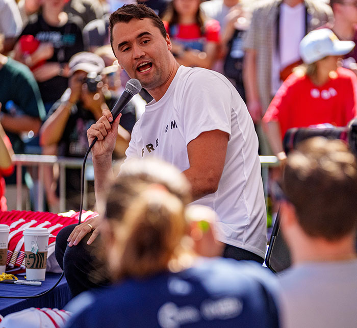 Charlie Kirk speaking into a microphone during an outdoor event with an audience in the background Charlie Kirk speaking into a microphone during an outdoor event with an audience in the background