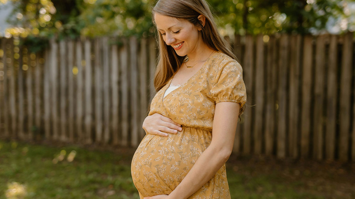 Pregnant mother in a yellow dress gently holding her belly outdoors, smiling with a wooden fence in the background.