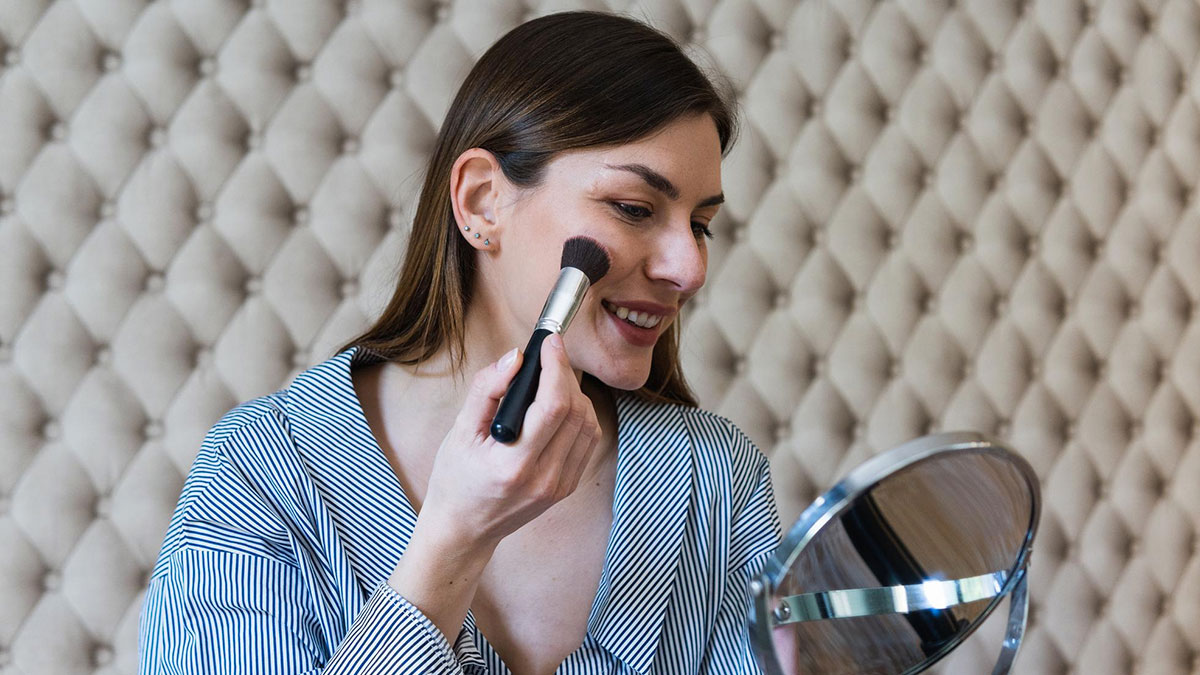 Woman applying makeup with a brush while smiling into a mirror, representing childish reasons ladies ditched a guy.