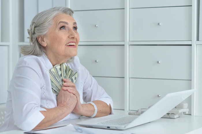 Elderly woman holding dollars with a hopeful expression, sitting at a desk near a laptop, symbolizing Christian rapture. Elderly woman holding dollars with a hopeful expression, sitting at a desk near a laptop, symbolizing Christian rapture.