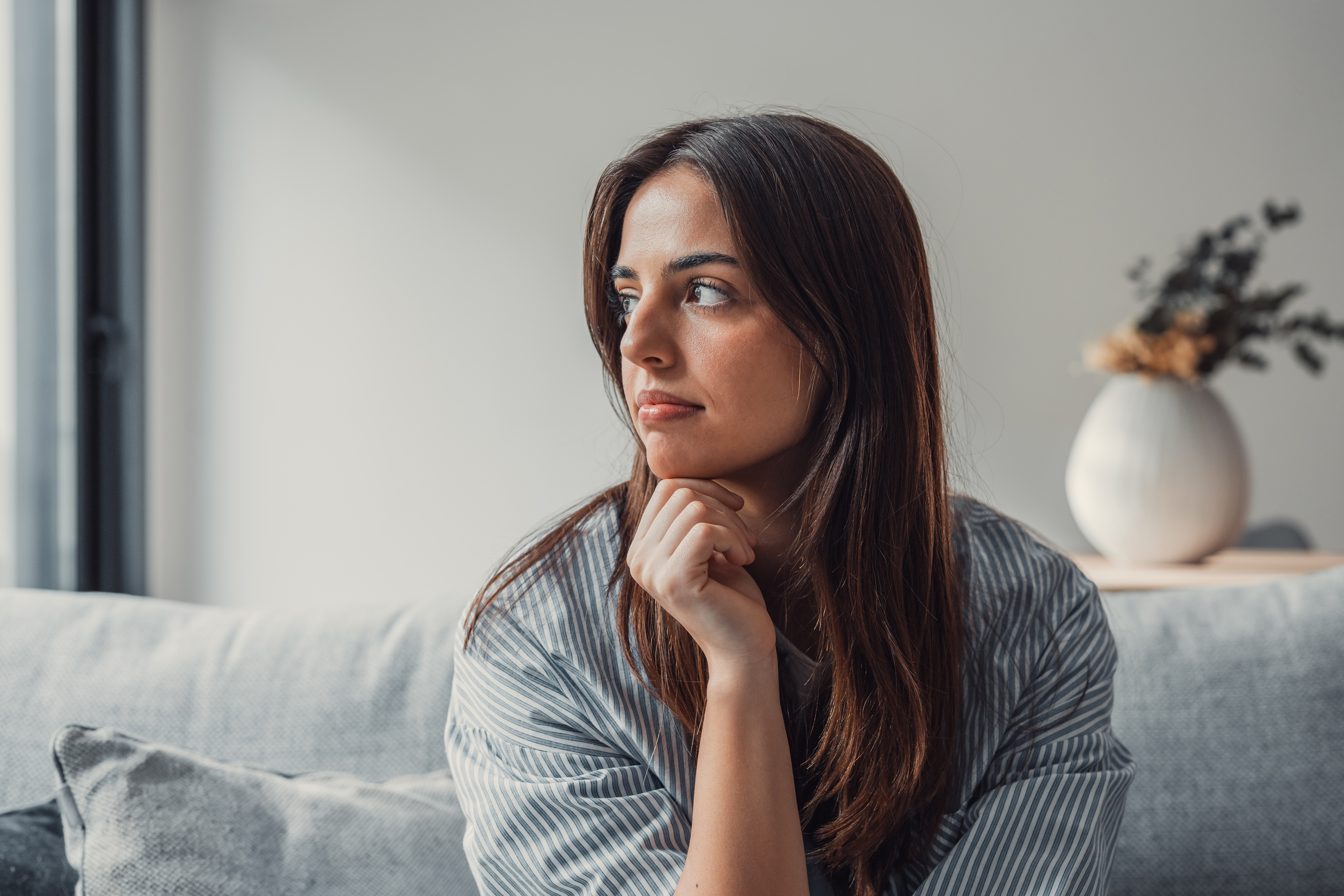 Woman sitting on sofa looking thoughtful, reflecting on boyfriend being affectionate with his mom, feeling conflicted. Woman sitting on sofa looking thoughtful, reflecting on boyfriend being affectionate with his mom, feeling conflicted.