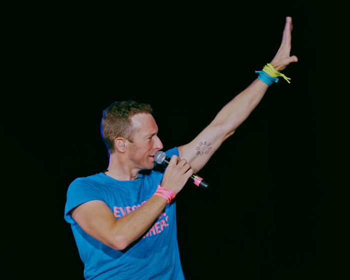 Coldplay’s Chris Martin speaking into a microphone on stage, raising his arm during a live concert moment. Coldplay’s Chris Martin speaking into a microphone on stage, raising his arm during a live concert moment.