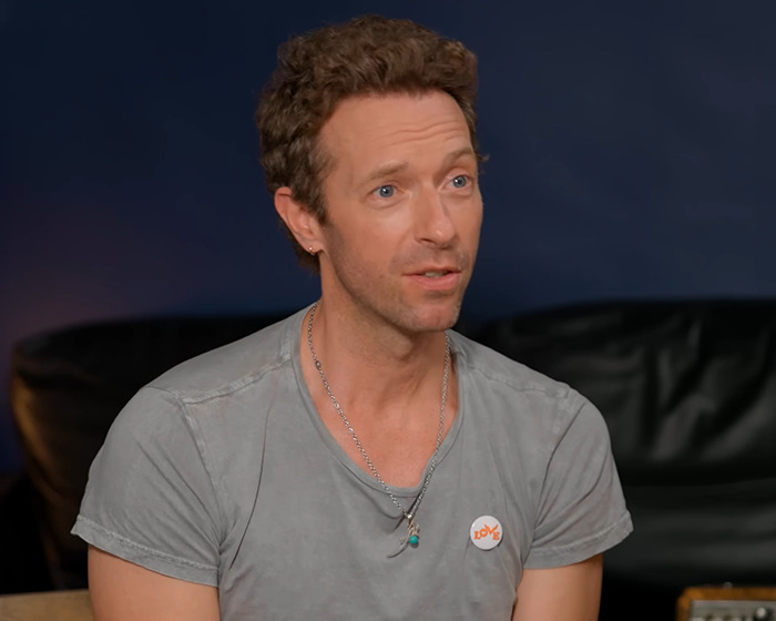 Chris Martin of Coldplay speaking during an interview, wearing a gray shirt with necklaces in a casual setting. Chris Martin of Coldplay speaking during an interview, wearing a gray shirt with necklaces in a casual setting.