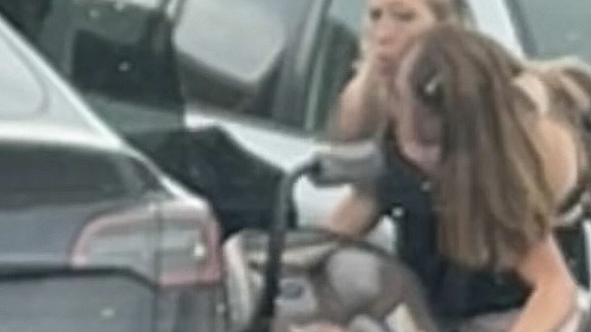 Conjoined twins Abby and Brittany at a gas station, focusing on a car with a blurred background.