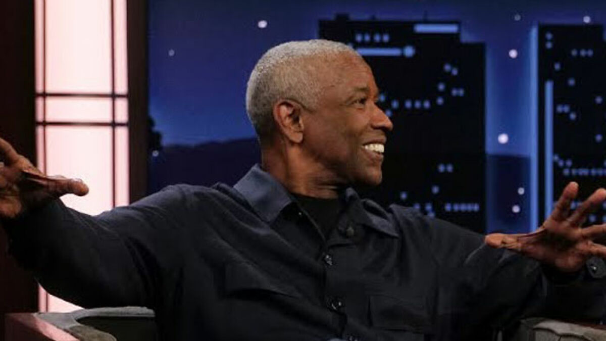 Denzel Washington smiling and gesturing during a nighttime talk show interview in a dark shirt against a citylight backdrop