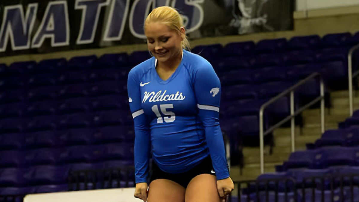 Cheerleader with baby belly wearing blue Wildcats uniform practicing in an empty indoor sports arena.