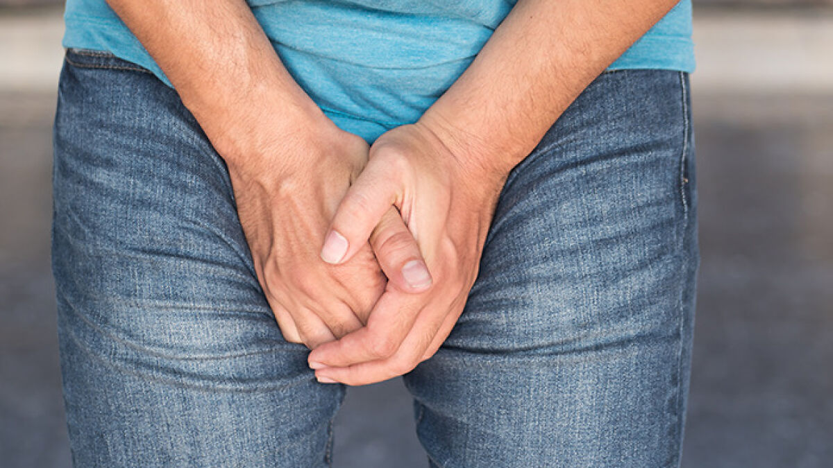 Man in jeans and blue shirt holding hands over groin area, relating to Scrotox cosmetic trend among men.