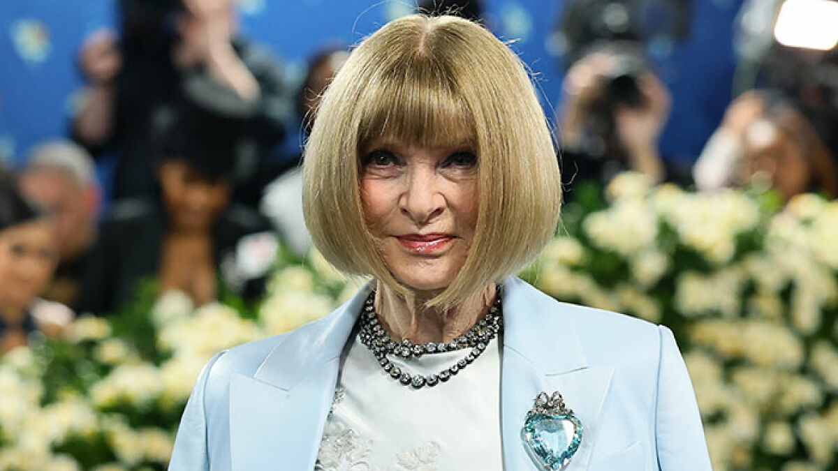 Anna Wintour in a light blue outfit and statement necklace, attending an event with photographers and flowers in the background.