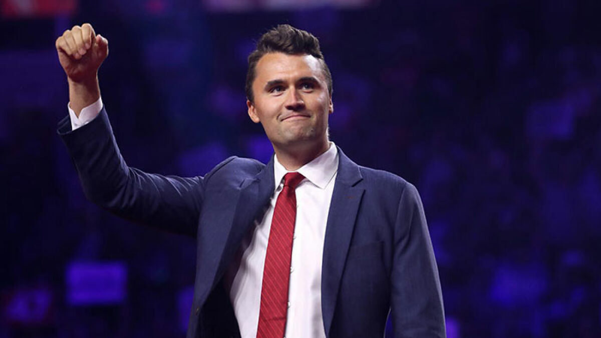 Charlie Kirk raising his fist on stage, wearing a navy suit and red tie, during a public speaking event.