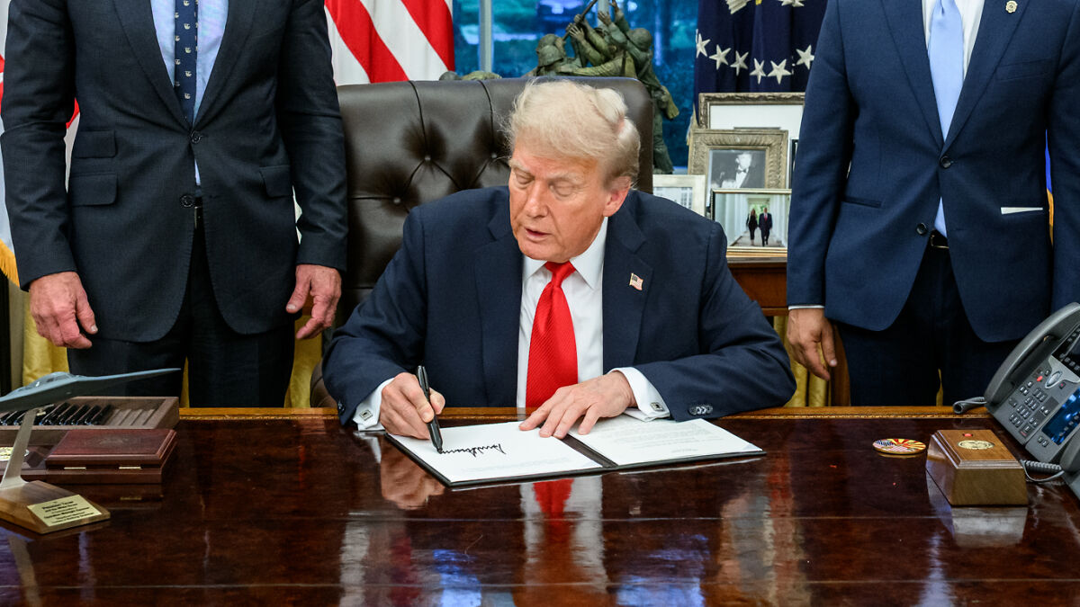 Man in a suit signing documents at a desk with two other men standing nearby, related to Charlie Kirk theory claims.
