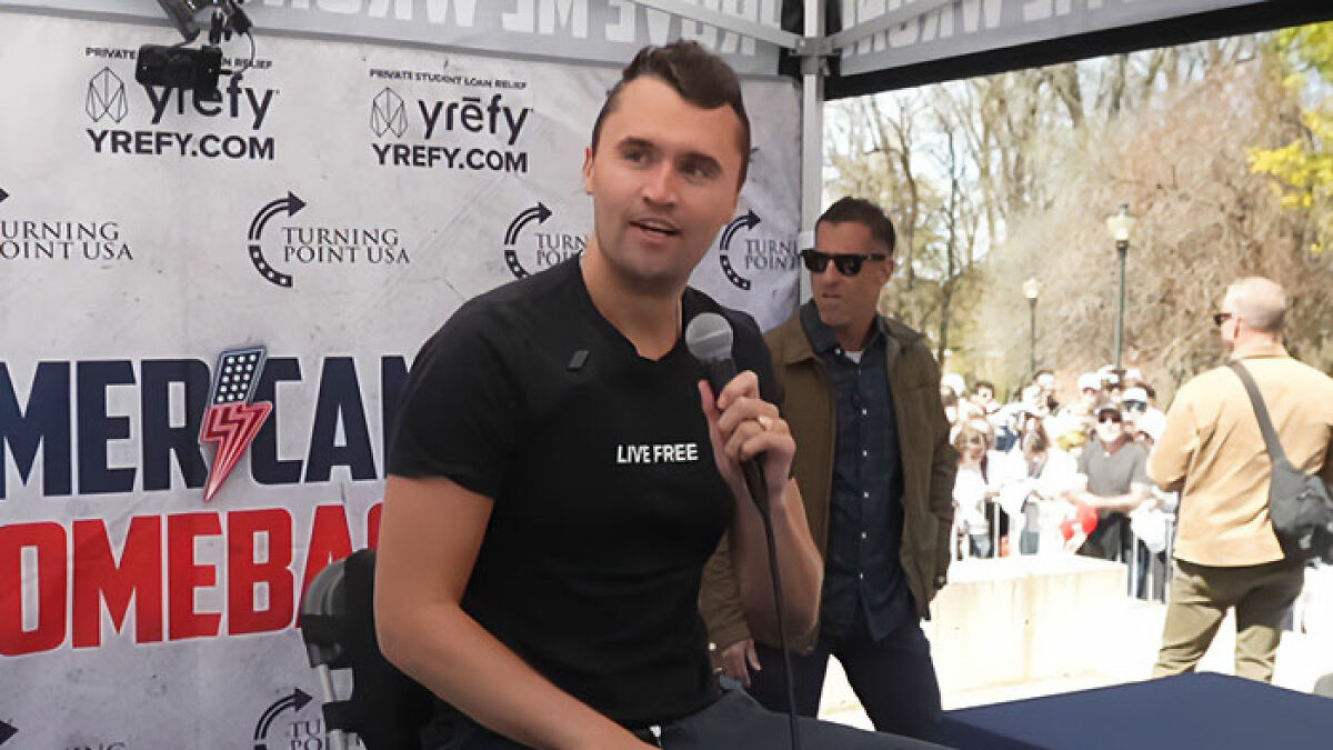 Man holding microphone speaking at Turning Point USA event with crowd outside, related to Charlie Kirk predicted passing.