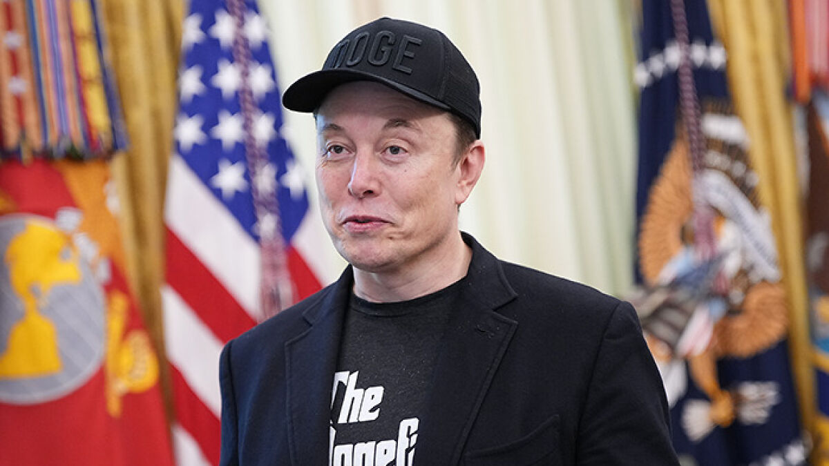 Elon Musk wearing a black cap and jacket delivering a strong one word response amidst US flags in the background