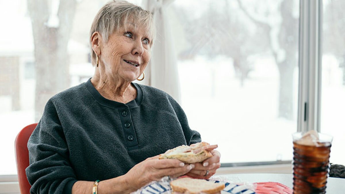 Elderly woman eating a sandwich indoors near a window, illustrating food related to increased dementia risk study.