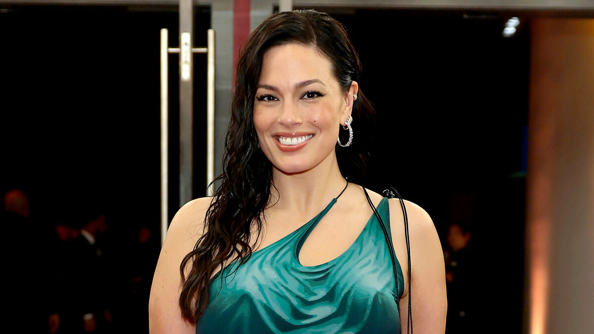Ashley Graham wearing a body-hugging emerald green dress, smiling with wet-look hair at an indoor event.