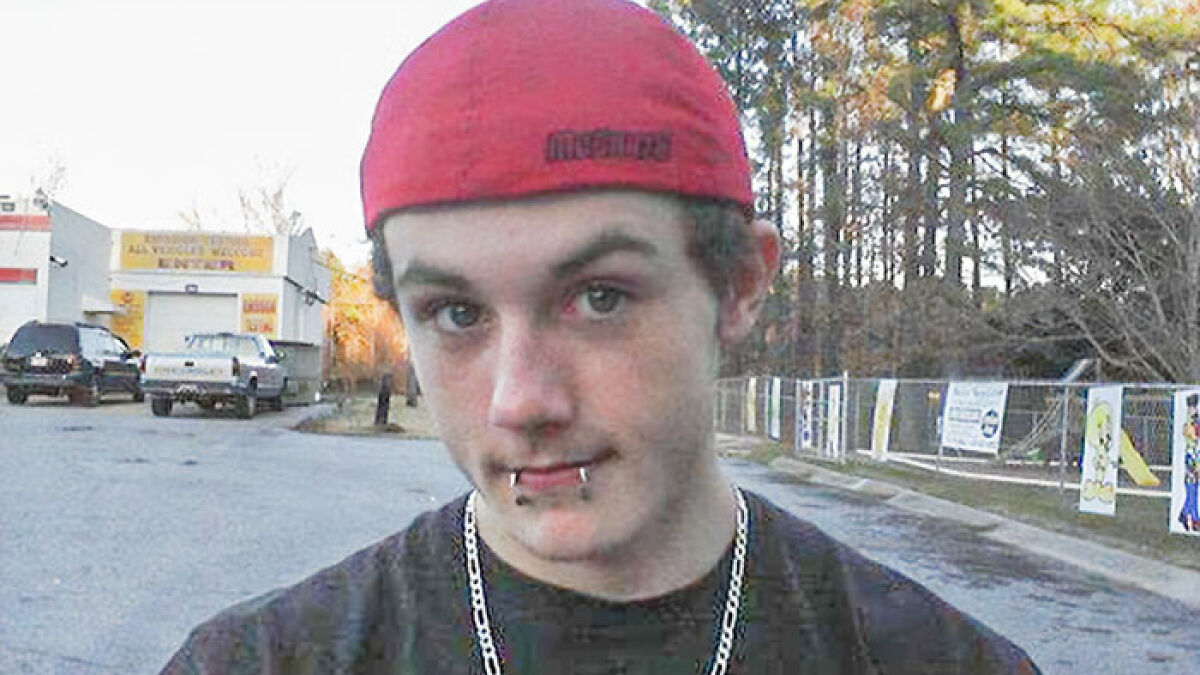 Teen boy with lip piercings wearing red cap outdoors near parking lot and fenced area with banners on trees background