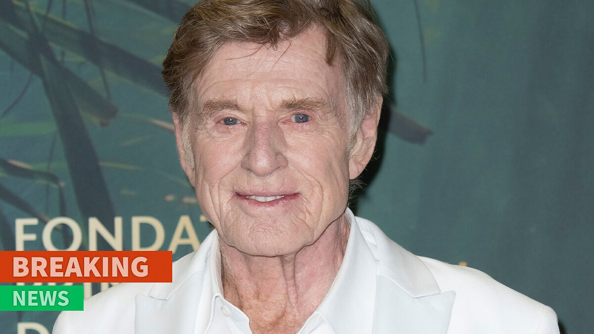 Robert Redford smiling at an event, wearing a white jacket against a green background with breaking news banner.
