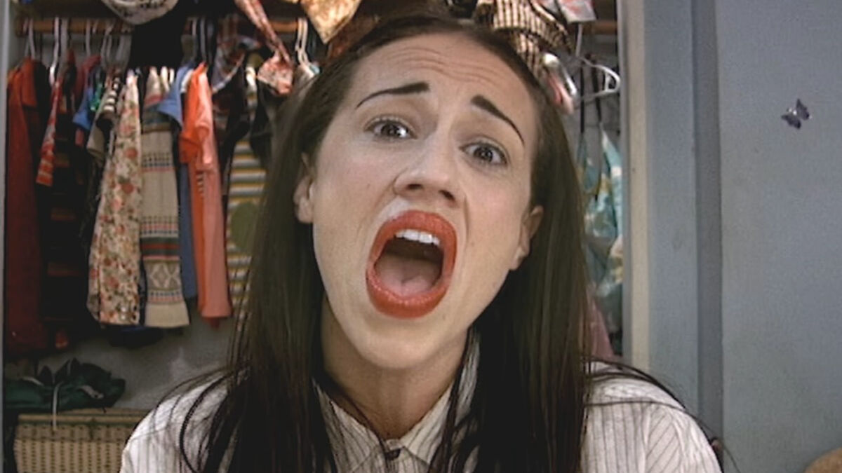 Woman with bold red lipstick singing or shouting in a room with clothes hanging behind, illustrating influencer career decline.