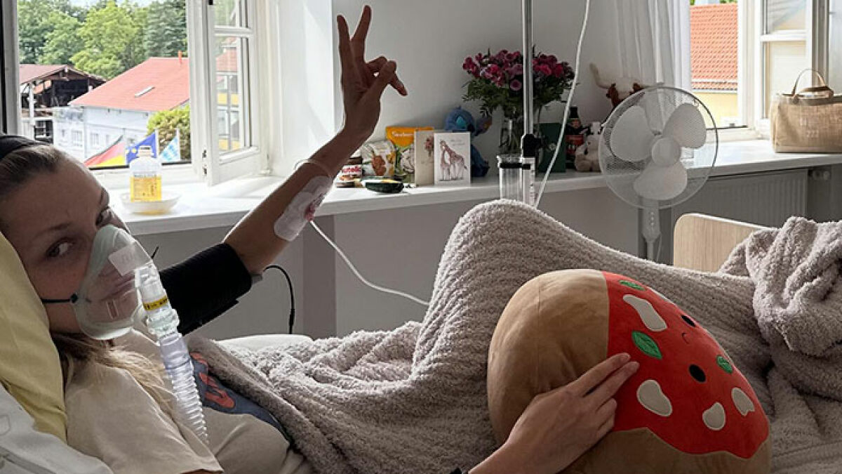 Bella Hadid in hospital bed wearing oxygen mask and IV, holding plush pillow, showing hand gesture during treatment.