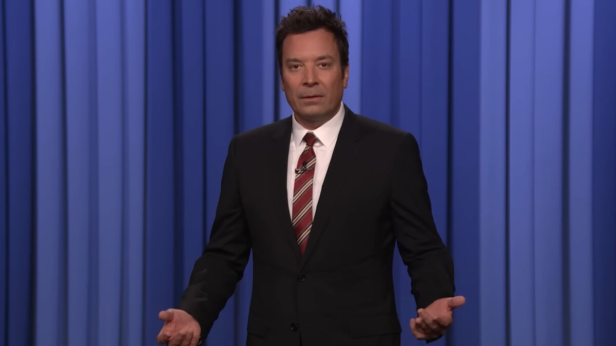 Jimmy Fallon in a dark suit and striped tie, speaking on stage with a blue curtain backdrop, addressing show suspension topics.