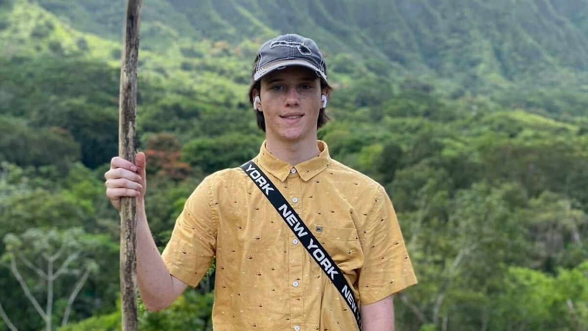 Young man wearing a yellow shirt and cap, holding a wooden stick, outdoors with green hills in the background.