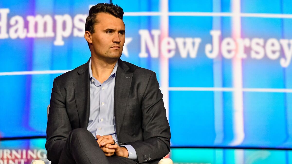 Charlie Kirk sitting on stage in a suit, with a blurred blue background mentioning New Jersey.