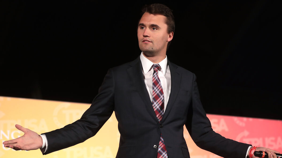 Charlie Kirk speaking at an event, wearing a suit and plaid tie, amid mass fury compared to historic funerals.