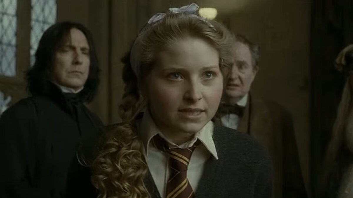 Harry Potter star in Hogwarts uniform speaking seriously with two men in the background in a dark room.