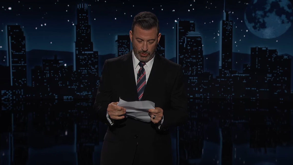 Jimmy Kimmel in a suit reading a tearful apology on stage about Charlie Kirk statements against a city skyline backdrop at night