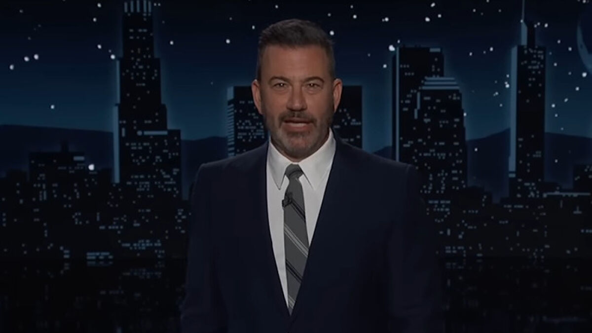 Jimmy Kimmel in a dark suit and striped tie delivering a monologue against a nighttime city skyline backdrop.