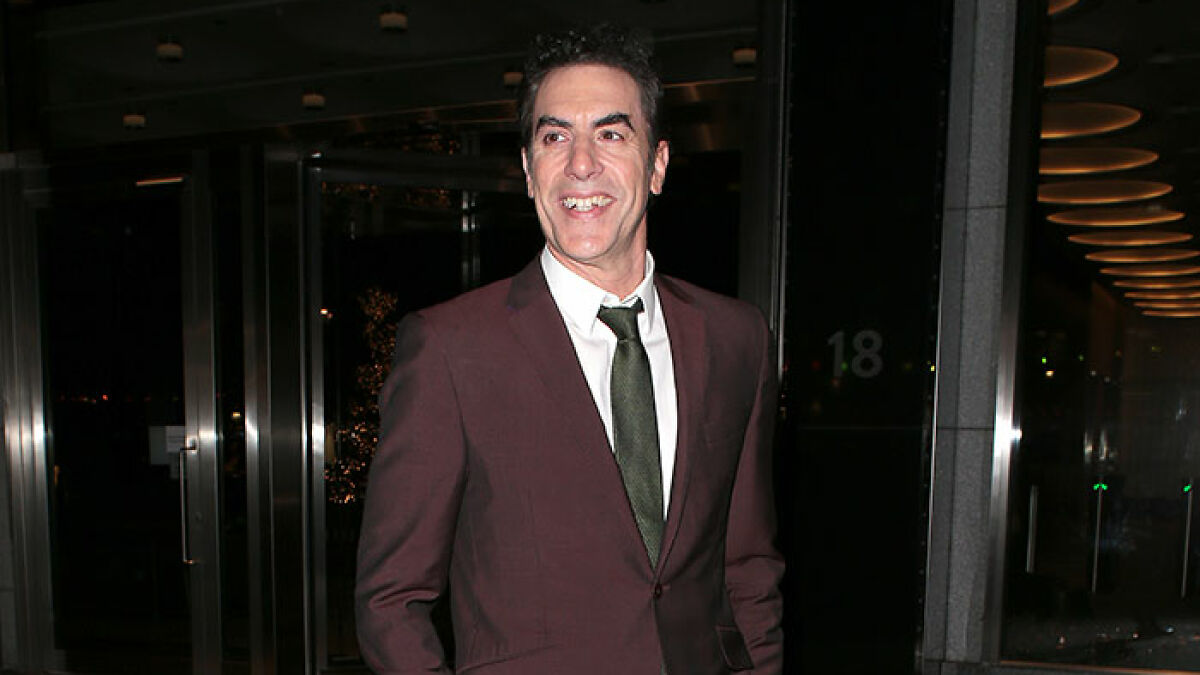 Sacha Baron Cohen smiling and wearing a maroon suit and green tie, standing outside a building at night.