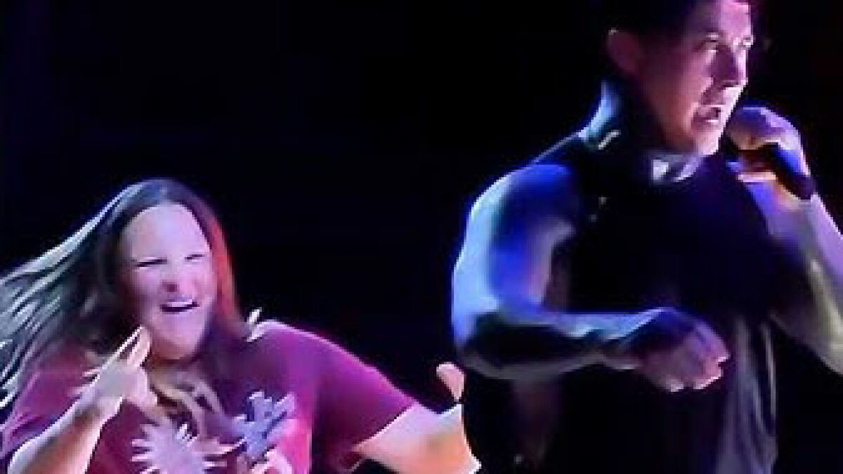 Ex-con rock star performing on stage as a woman who rushed the stage reacts in the background.