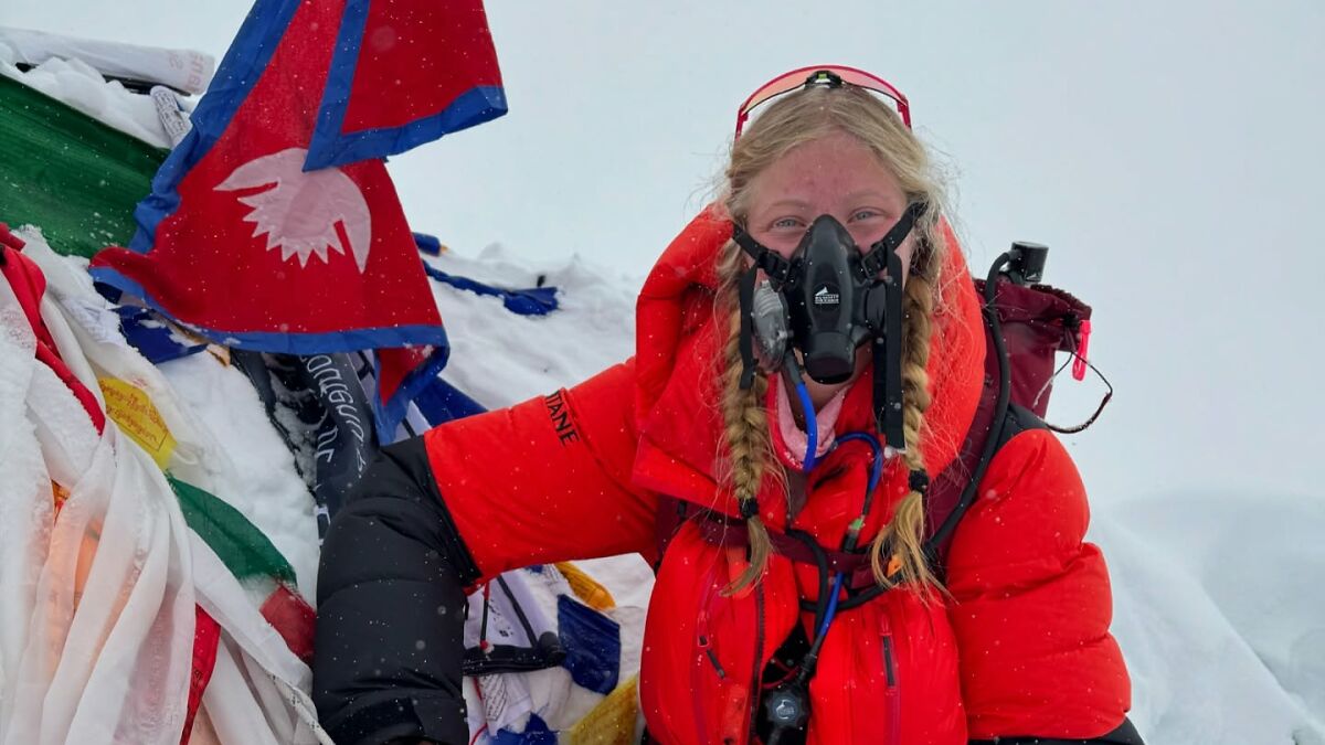 17-year-old climber wearing oxygen mask and red jacket on Everest amid snow with Nepalese flag in the background.