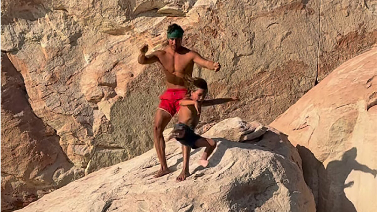 Man wearing red shorts and headband appearing to push young boy off a cliff amid viral tough love outrage.