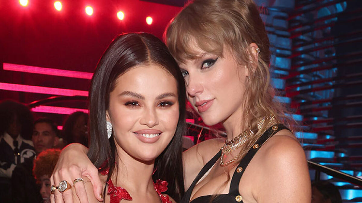 Taylor Swift and Selena Gomez smiling together at an event with dramatic red and blue lighting in the background.