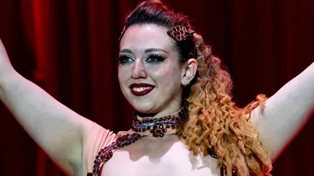 Trapeze artist with elaborate stage makeup and costume performing under red circus curtains during stunt.