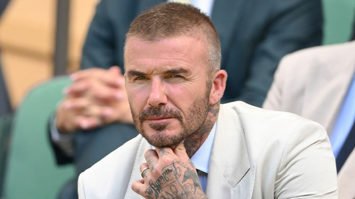 David Beckham sitting thoughtfully with hand on chin, showing tattoos and a serious expression in a light suit.