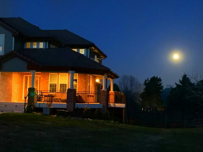 A warmly lit house at night under a bright full moon, evoking a quiet scene after bizarre phone calls and messages.