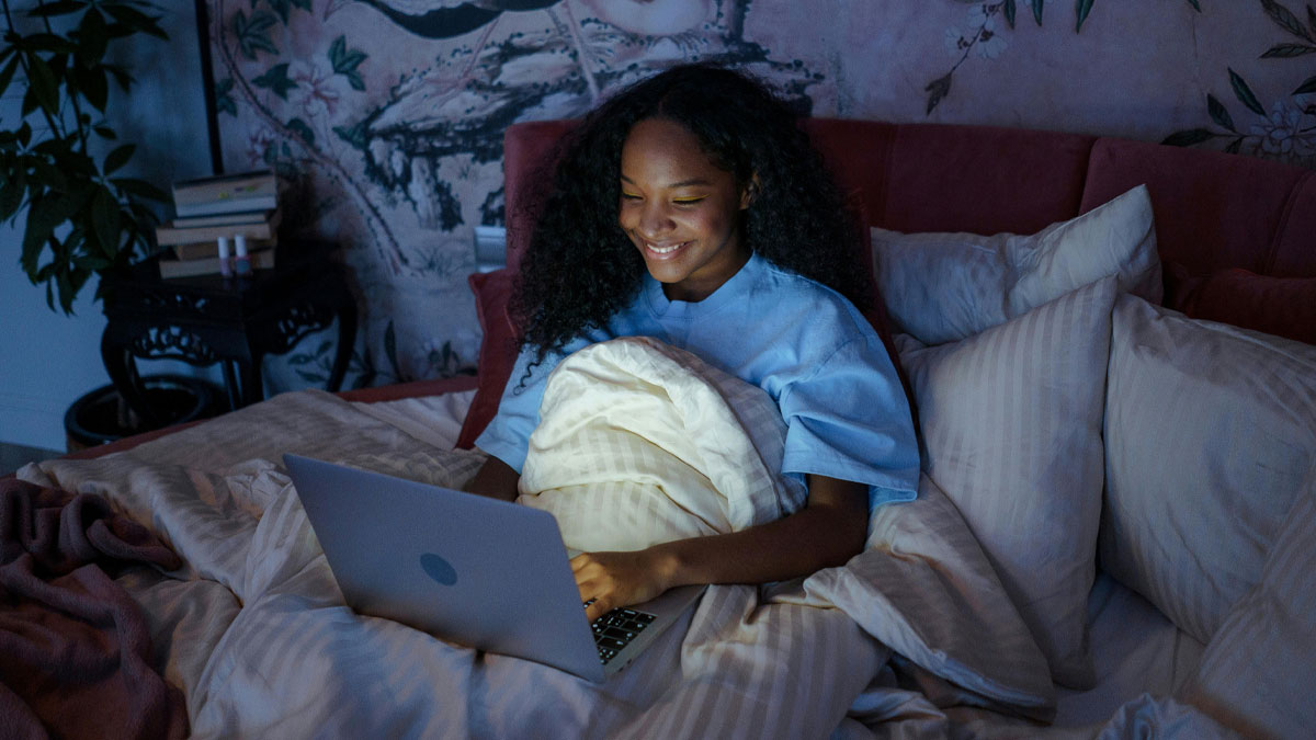 Young woman in bed smiling while reading unbelievable and horrifying things people swear they saw on a laptop at night