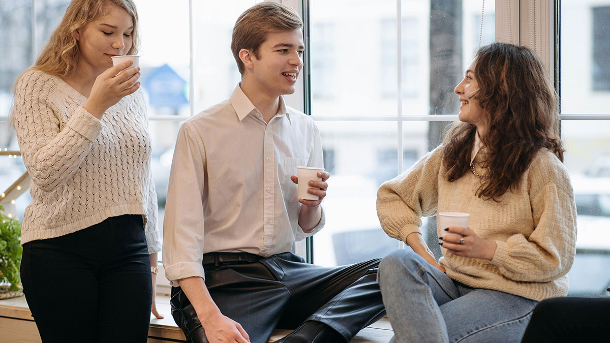Young man holding a cup looking uncomfortable while two women chat and drink coffee by a large window in a casual setting