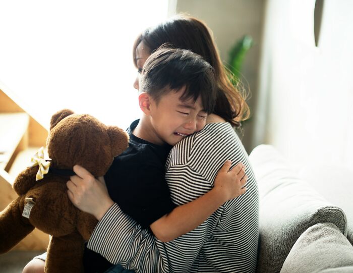 Child crying while hugging mother holding a teddy bear, illustrating parenting hacks to save sanity at home.