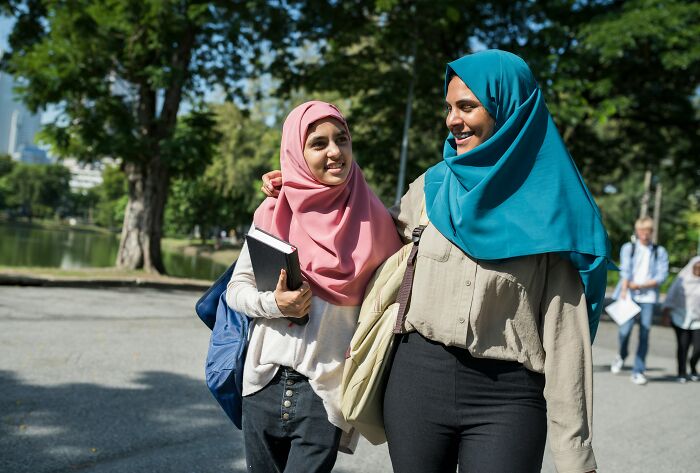 Two female students wearing hijabs, walking and smiling outdoors on a school campus, carrying books and bags.